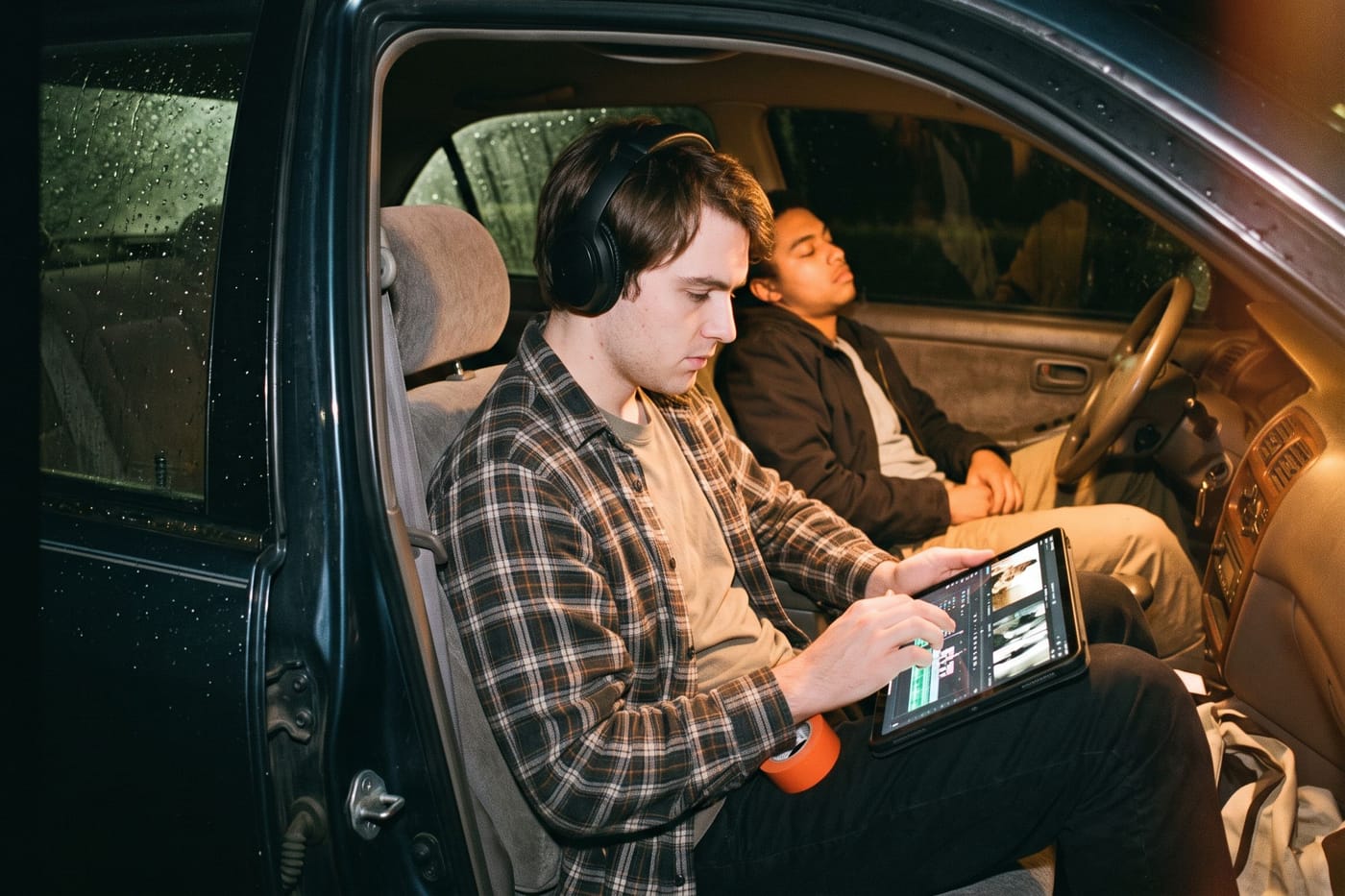 An editor cutting on a tablet in a parked car at night while a crew member sleeps in the back seat